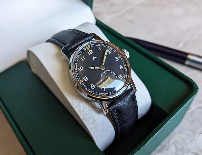May include: A black leather wristwatch with a silver case and a black face. The watch has white numerals and a white second hand. The watch is in a green box.
