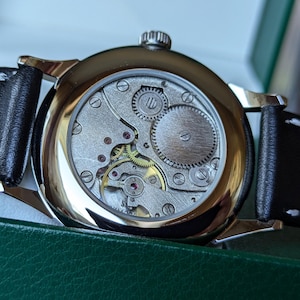May include: A silver watch with a black leather band. The back of the watch is open, revealing the intricate mechanical workings of the movement.