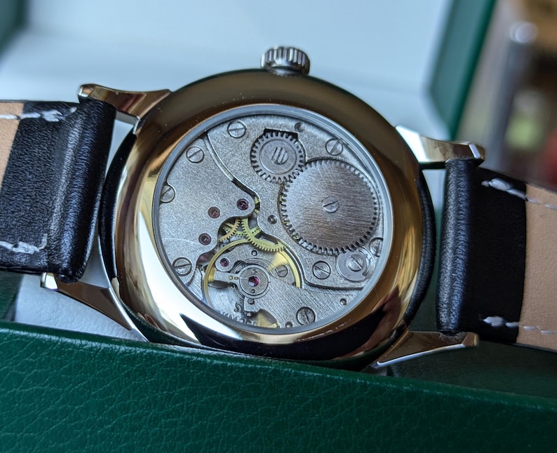 May include: A silver watch with a black leather band. The back of the watch is open, revealing the intricate mechanical workings of the movement.