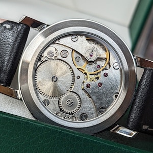 May include: A silver watch with a black leather band. The watch face is open, revealing the intricate inner workings of the watch mechanism.