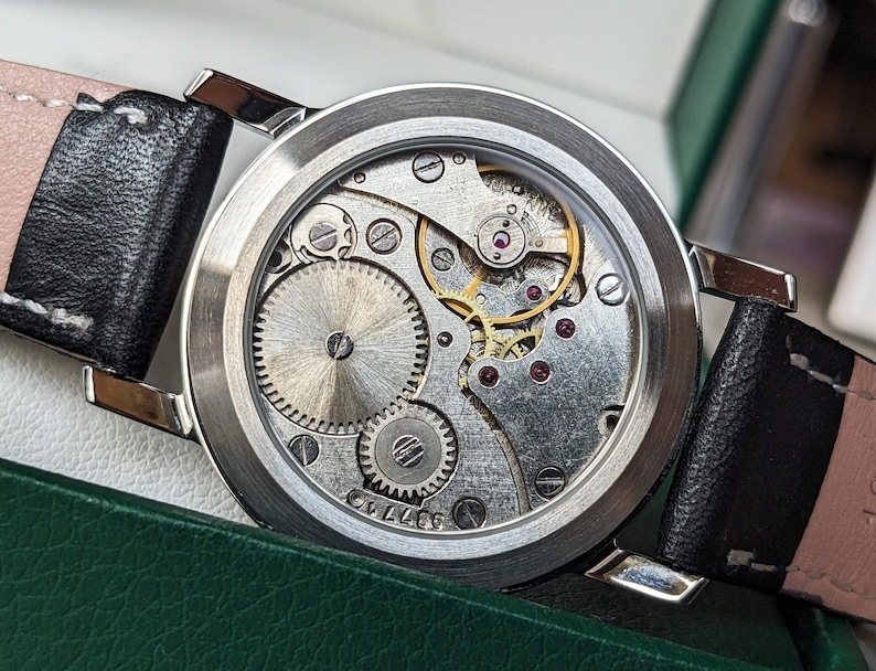 May include: A silver watch with a black leather band. The watch face is open, revealing the intricate inner workings of the watch mechanism.