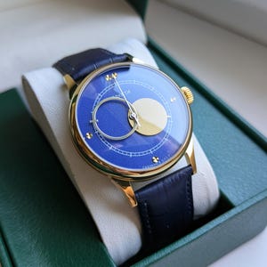 May include: A gold-toned wristwatch with a blue face and a black leather band. The watch face features a gold crescent and a circular design. The watch is presented in a green box with a white interior.