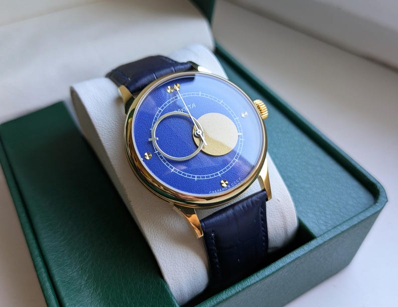 May include: A gold-toned wristwatch with a blue face and a black leather band. The watch face features a gold crescent and a circular design. The watch is presented in a green box with a white interior.