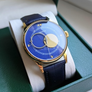 May include: A gold-toned wristwatch with a blue face and a black leather band. The watch face features a gold-colored crescent and a circular design. The watch is presented in a green box with a white interior.