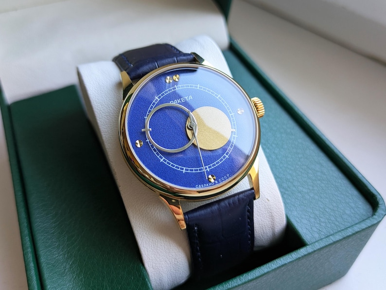 May include: A gold-toned wristwatch with a blue face and a black leather band. The watch face features a gold-colored crescent and a circular design. The watch is presented in a green box with a white interior.