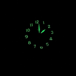 May include: A black clock face with green luminous numbers and hands. The time is 12:00.