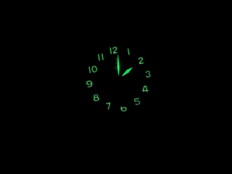 May include: A black clock face with green luminous numbers and hands. The time is 12:00.