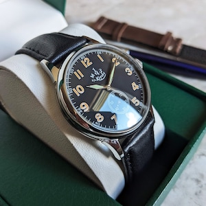May include: A black leather wristwatch with a silver case and a black face. The watch has white numerals and a small second hand. The watch is in a green box with a white interior.