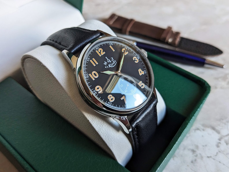 May include: A black leather wristwatch with a silver case and a black face. The watch has white numerals and a small second hand. The watch is in a green box with a white interior.