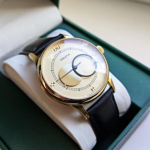 May include: A gold-tone wristwatch with a black leather band. The watch face has a white background with black markings and the word "PAKETA" printed on it.