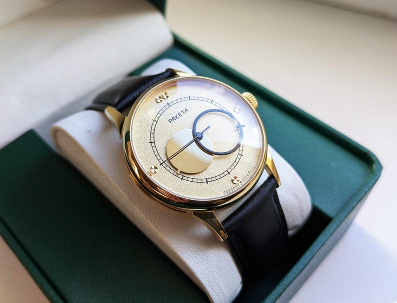 May include: A gold-tone wristwatch with a black leather band. The watch face has a white background with black markings and the word "PAKETA" printed on it.