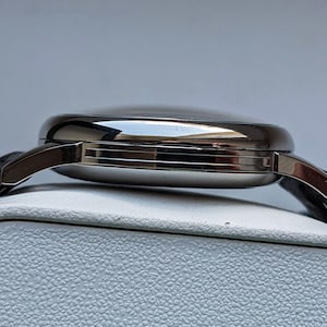 May include: Side view of a silver wristwatch with a black leather band. The watch has a round face and a polished finish.