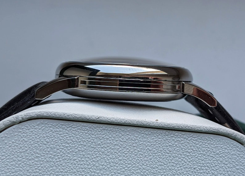 May include: Side view of a silver wristwatch with a black leather band. The watch has a round face and a polished finish.