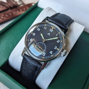 May include: A black leather wristwatch with a silver case and a black face. The watch has white numerals and a small second hand. The watch is in a green box.