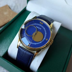 May include: A gold-toned Raketa watch with a blue face and a blue leather band. The watch face features a unique design with a gold-colored circle and a smaller, silver-colored circle. The watch is presented in a green box.