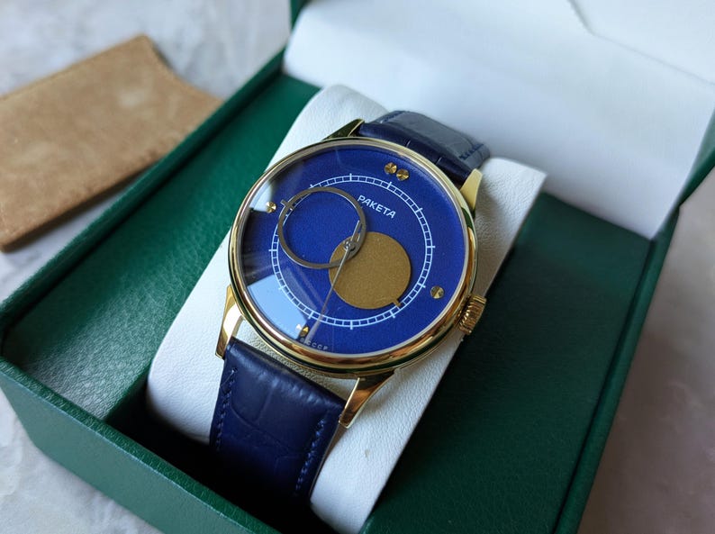 May include: A gold-toned Raketa watch with a blue face and a blue leather band. The watch face features a unique design with a gold-colored circle and a smaller, silver-colored circle. The watch is presented in a green box.