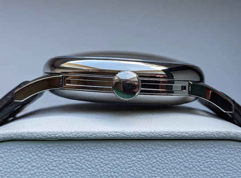 May include: A silver wristwatch with a round face and a black leather band. The watch has a silver crown on the right side of the case.