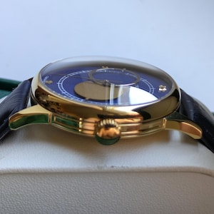 May include: A luxury wristwatch with a gold-tone case and a dark blue dial. The watch features a black leather strap and a domed crystal. The dial has gold-tone accents and a moon phase indicator. The watch is displayed on a white surface.