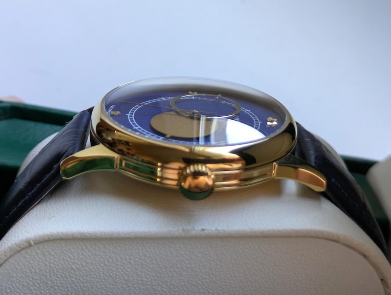 May include: A luxury wristwatch with a gold-tone case and a dark blue dial. The watch features a black leather strap and a domed crystal. The dial has gold-tone accents and a moon phase indicator. The watch is displayed on a white surface.