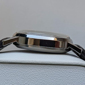 May include: A silver wristwatch with a black leather band. The watch is on a white display stand.