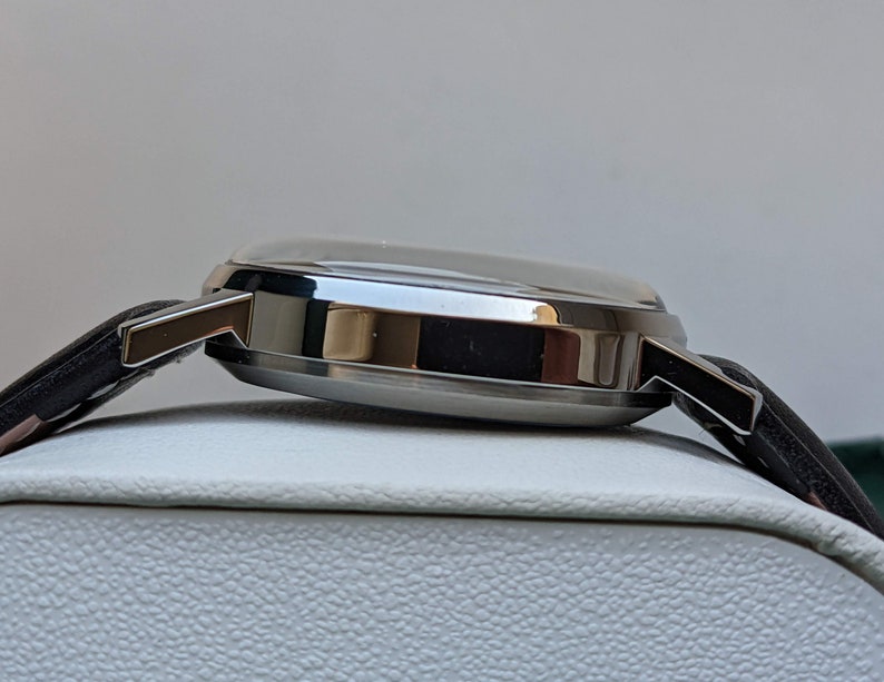 May include: A silver wristwatch with a black leather band. The watch is on a white display stand.