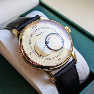 May include: A gold-tone wristwatch with a black leather band. The watch face is white with a gold-tone circle in the center and the word "PAKETA" printed on the face.