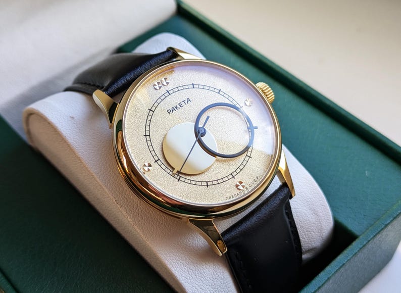 May include: A gold-tone wristwatch with a black leather band. The watch face is white with a gold-tone circle in the center and the word "PAKETA" printed on the face.