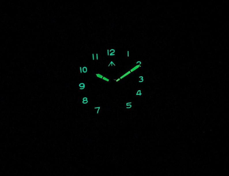 May include: A black watch face with green luminous hour markers and hands. The watch face shows the numbers 1 through 12.