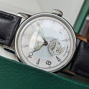 May include: A silver wristwatch with a white face and black leather band. The watch face has black Roman numeral hour markers and a small subdial with a compass design. The watch face also has the text "Сделано в СССР" and "143 Чистополь" printed on it.