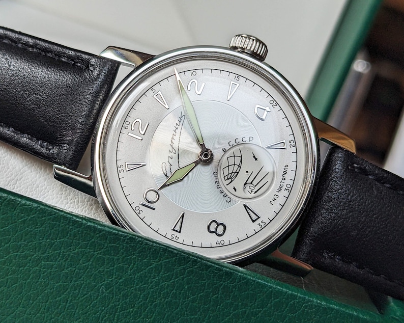 May include: A silver wristwatch with a white face and black leather band. The watch face has black Roman numeral hour markers and a small subdial with a compass design. The watch face also has the text "Сделано в СССР" and "143 Чистополь" printed on it.