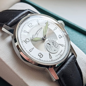 May include: A silver wristwatch with a white face and black leather band. The watch face has black Roman numeral hour markers, a black second hand, and a small subdial with a moon phase indicator. The watch face also has the text "Спутник" and "СССР" printed on it.