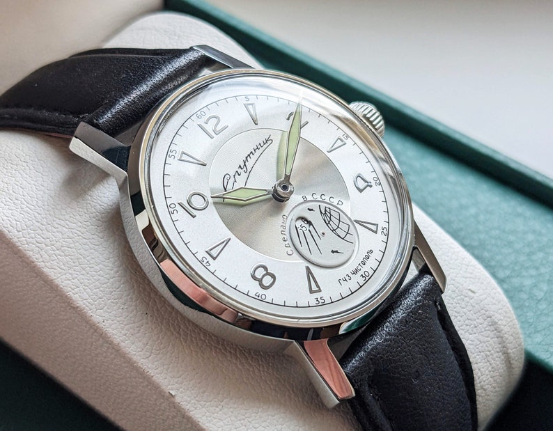 May include: A silver wristwatch with a white face and black leather band. The watch face has black Roman numeral hour markers, a black second hand, and a small subdial with a moon phase indicator. The watch face also has the text "Спутник" and "СССР" printed on it.