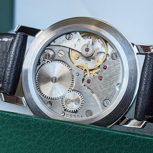 May include: A silver watch with a black leather band and a clear case back revealing the inner workings of the watch mechanism.