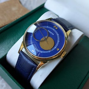 May include: A gold-toned wristwatch with a blue face and a navy blue leather band. The watch face features the word "RAKETA" and a gold-colored circle and crescent design. The watch is presented in a green box with a tan suede pouch.