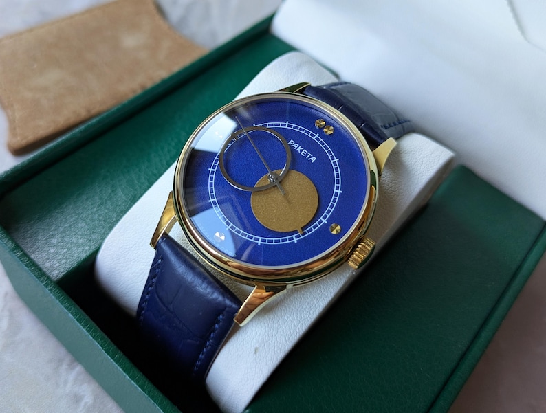 May include: A gold-toned wristwatch with a blue face and a navy blue leather band. The watch face features the word "RAKETA" and a gold-colored circle and crescent design. The watch is presented in a green box with a tan suede pouch.
