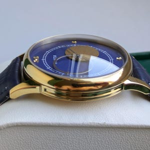 May include: A gold-toned wristwatch with a blue face and a dark blue leather band. The watch face has gold accents and a clear crystal cover. The watch is displayed on a white surface.
