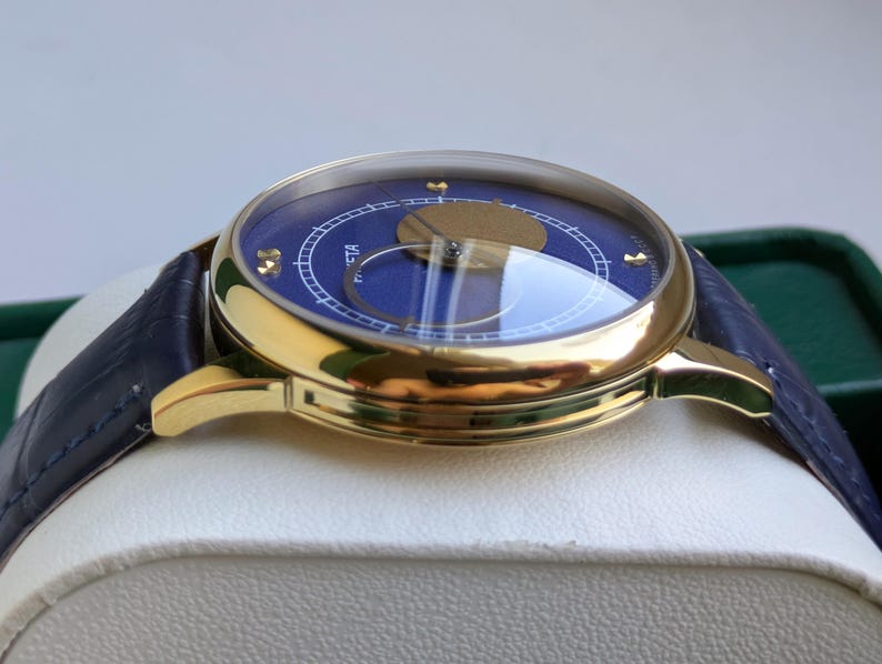May include: A gold-toned wristwatch with a blue face and a dark blue leather band. The watch face has gold accents and a clear crystal cover. The watch is displayed on a white surface.