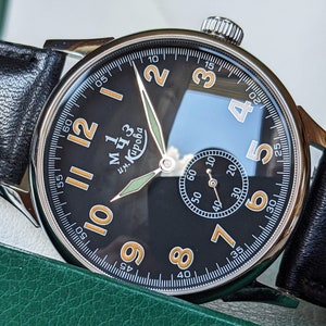 May include: A black wristwatch with a white face and orange numbers. The watch has a black leather band and a silver case. The watch face has the text "им. Кирова" in Cyrillic script.