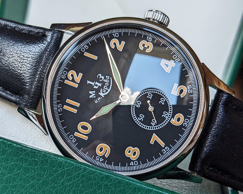 May include: A black wristwatch with a white face and orange numbers. The watch has a black leather band and a silver case. The watch face has the text "им. Кирова" in Cyrillic script.