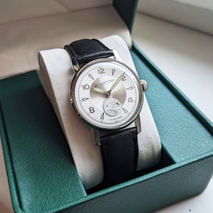 May include: A silver wristwatch with a white face and black leather band. The watch face has black Roman numeral hour markers and a green second hand. The watch is in a green box.