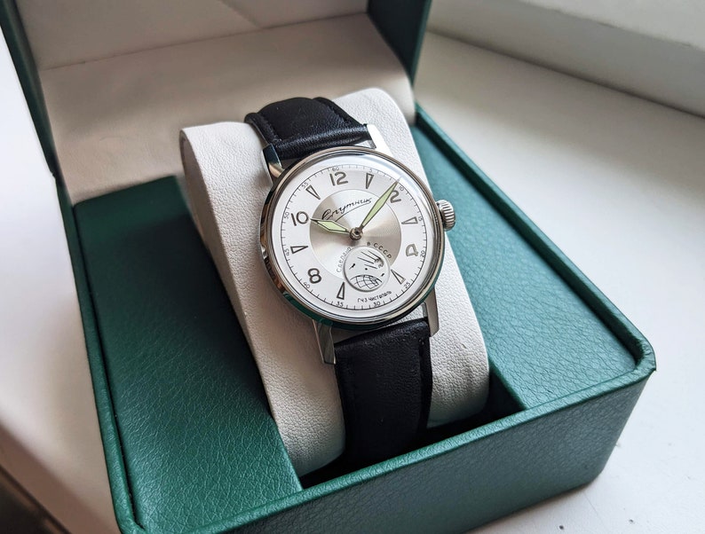 May include: A silver wristwatch with a white face and black leather band. The watch face has black Roman numeral hour markers and a green second hand. The watch is in a green box.