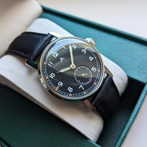 May include: A black-faced wristwatch with white numerals and a black leather band. The watch is in a green box.