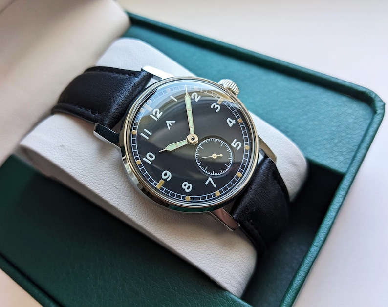 May include: A black-faced wristwatch with white numerals and a black leather band. The watch is in a green box.