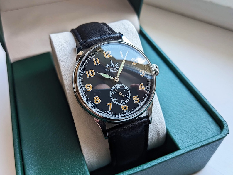May include: A black leather wristwatch with a silver case and a black face. The watch has white numerals and a small second hand. The watch face also has the text "M.Ч.З. им. Кирова" in gold lettering.