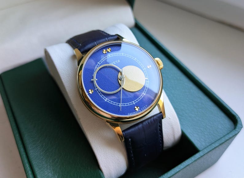 May include: A gold-toned wristwatch with a blue face and a gold-colored crescent and circle design. The watch has a black leather band and is displayed in a green box. The watch face has the word "Raketa" on it.