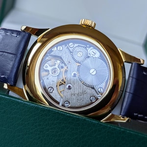 May include: A gold-toned wristwatch with a transparent back, showcasing the intricate internal mechanisms. The watch has a dark blue leather strap and a gold crown. The watch is set against a green background.
