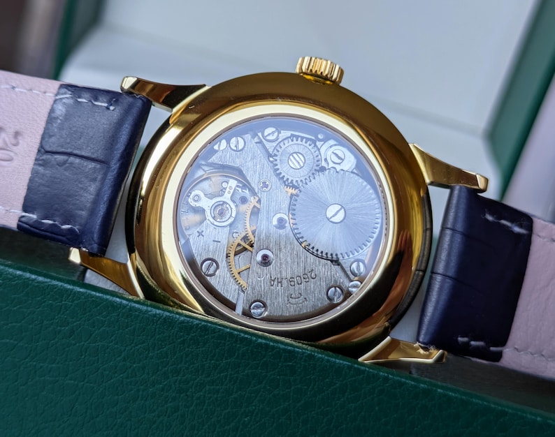 May include: A gold-toned wristwatch with a transparent back, showcasing the intricate internal mechanisms. The watch has a dark blue leather strap and a gold crown. The watch is set against a green background.