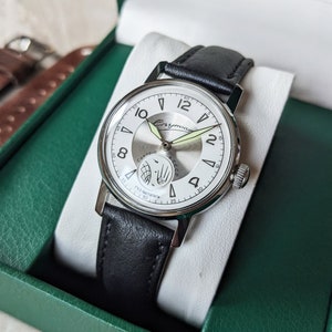 May include: A silver wristwatch with a white face and black leather band. The watch face has black Roman numeral hour markers and a small subdial at the 3 o'clock position. The watch is in a green box with a brown leather strap.