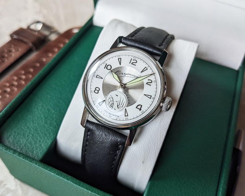 May include: A silver wristwatch with a white face and black leather band. The watch face has black Roman numeral hour markers and a small subdial at the 3 o'clock position. The watch is in a green box with a brown leather strap.