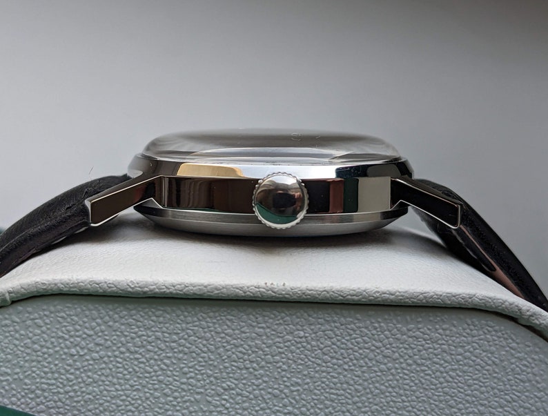 May include: A silver wristwatch with a black leather band. The watch has a round face and a silver case. The watch is on a white surface.